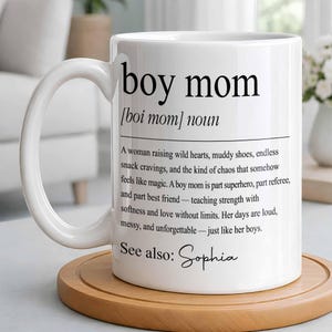 May include: White ceramic mug with the words "boy mom" in black, along with a definition. The mug has a curved handle and sits on a round wooden coaster. The text includes the name "Sophia".