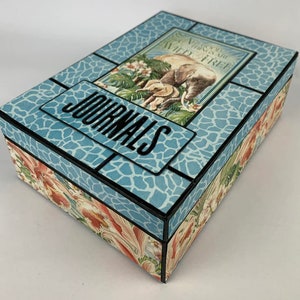 May include: A blue box with a black border and a floral pattern on the bottom. The top of the box has a blue and white patterned design with a black border and the word "JOURNALS" in black letters. The box also has a picture of two elephants and the text "Things are Wild & Free".