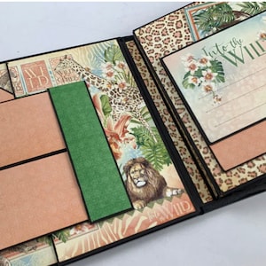 Puede incluir: &Aacute;lbum de recortes abierto con tem&aacute;tica de jungla. Las p&aacute;ginas presentan ilustraciones de un le&oacute;n, estampado de leopardo y dise&ntilde;os florales. El texto "Into the Wild" es visible en una p&aacute;gina. Incluye dos etiquetas marrones con ojales negros y un acento verde.