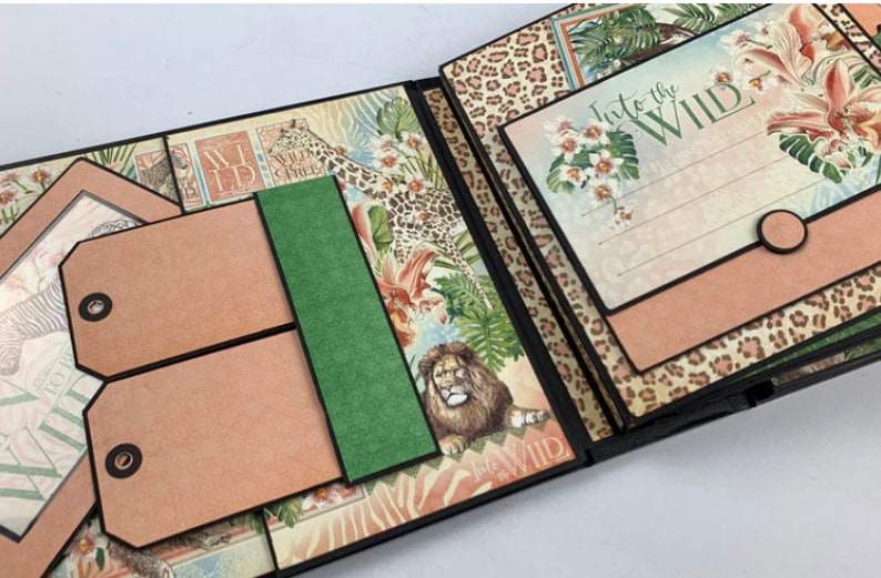 Puede incluir: &Aacute;lbum de recortes abierto con tem&aacute;tica de jungla. Las p&aacute;ginas presentan ilustraciones de un le&oacute;n, estampado de leopardo y dise&ntilde;os florales. El texto "Into the Wild" es visible en una p&aacute;gina. Incluye dos etiquetas marrones con ojales negros y un acento verde.