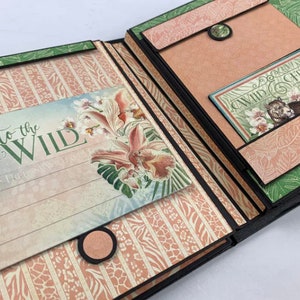 Puede incluir: &Aacute;lbum de recortes abierto con tem&aacute;tica de jungla. Las p&aacute;ginas presentan patrones florales y de animales en tonos rosa, verde y beige. El texto "Into the Wild" es visible en una p&aacute;gina y "Wild & Free" en otra. Elementos decorativos con ilustraciones florales.