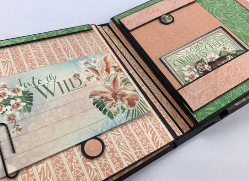 Puede incluir: &Aacute;lbum de recortes abierto con tem&aacute;tica de jungla. Las p&aacute;ginas presentan patrones florales y de animales en tonos rosa, verde y beige. El texto "Into the Wild" es visible en una p&aacute;gina y "Wild & Free" en otra. Elementos decorativos con ilustraciones florales.