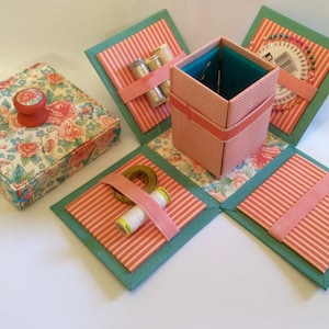 May include: A pink and green fabric box with a floral pattern and a pink ribbon. The box is open and has compartments for sewing supplies, including needles, thread, and a tape measure.