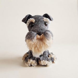 Könnte beinhalten: Eine kleine, handgefertigte Häkelfigur eines Schnauzers. Das Plüschtier besteht aus dunkelgrauem Garn für Körper und Kopf, mit helleren, beigefarbenen, strukturierten Garnakzenten an Schnauze, Augenbrauen und Pfoten. Es hat zwei schwarze Perlenaugen.