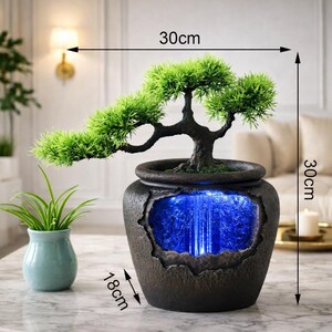 May include: A decorative bonsai tree in a dark brown pot with a blue illuminated waterfall feature. The pot is 30 cm tall and 30 cm wide, with a base of 18 cm. A small, light blue vase with a green plant is to the left.