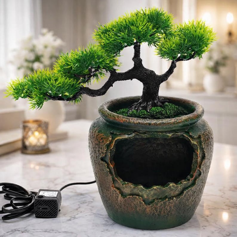 May include: A decorative bonsai tree in a weathered green and brown ceramic pot. The tree has vibrant green foliage and a dark, twisted trunk. A small black pump with a power cord is connected to the pot.