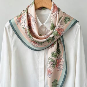 May include: A silk scarf with a floral pattern. The scarf features a light pink background with large pink and peach flowers and green leaves. The border is a muted green. The scarf is draped over a white collared shirt.