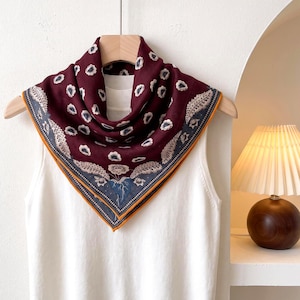 Può includere: Un foulard quadrato color bordeaux intenso con un motivo ripetitivo di piccoli fiori bianchi. Il foulard presenta un bordo ampio con dettagliate illustrazioni botaniche blu e beige e un sottile profilo giallo senape, drappeggiato su una canotta.
