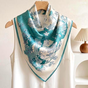 May include: A square scarf featuring a teal and white botanical print with illustrations of rabbits nestled among trees and flowers. The scarf is draped over a wooden hanger and layered over a sleeveless white top, showcasing a smooth texture.