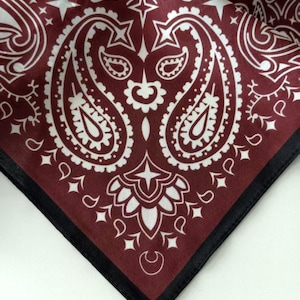 May include: A square fabric bandana featuring a deep burgundy background with a white paisley pattern. The design includes intricate teardrop shapes, floral motifs, and small star accents. The edges are finished with a solid black stitched border.