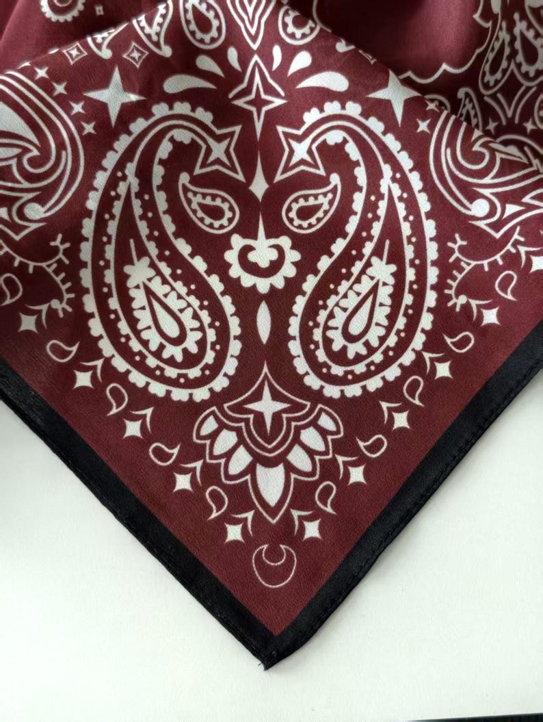 May include: A square fabric bandana featuring a deep burgundy background with a white paisley pattern. The design includes intricate teardrop shapes, floral motifs, and small star accents. The edges are finished with a solid black stitched border.