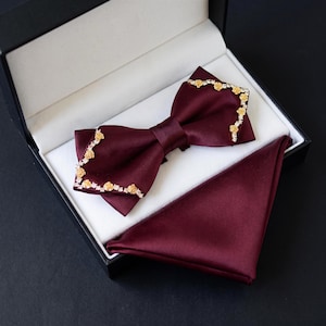 May include: A deep burgundy satin bow tie and a matching pocket square set. The bow tie features gold-toned floral embellishments and clear crystals along the outer edges. The set is presented inside a black rectangular box with a white interior.