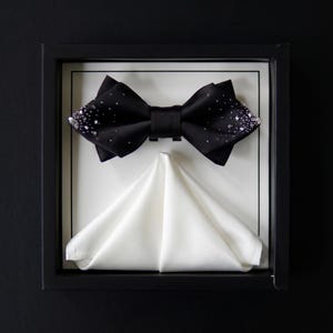 May include: A black satin bow tie featuring scattered clear crystal embellishments on the outer edges, paired with a folded white pocket square. Both items are presented inside a black display box with a white interior backing and black border.