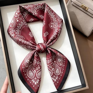 May include: A square scarf in a deep burgundy red color featuring a white paisley pattern. The scarf is tied in a knot at the center, displaying symmetrical folded ends with a solid black border along the edges of the fabric design.