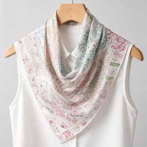 Op de afbeelding: Een vierkante zijden sjaal met een bloemenpatroon in pastelkleuren roze, groen en crème. De sjaal is gedrapeerd over een witte mouwloze top en een houten hanger. Het ontwerp bevat botanische en sierlijke elementen.