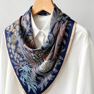 May include: A square silk scarf with a navy blue background and a botanical print. The scarf features a detailed design of leaves and flowers in shades of pink, green, and beige, with a gold border. The scarf is draped over a white collared shirt.
