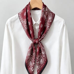 May include: A square scarf featuring a deep burgundy background with a white paisley and floral pattern. The scarf is tied in a knot at the center and is draped over a crisp white collared button-down shirt hanging on a wooden hanger.