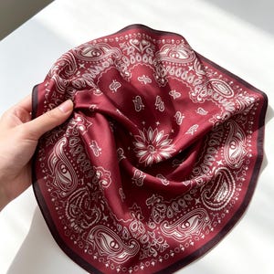 May include: A square scarf in a deep burgundy red color featuring a white printed paisley pattern with intricate swirling motifs and a central floral design. The fabric has a smooth, satin-like finish with a dark border along the edges.