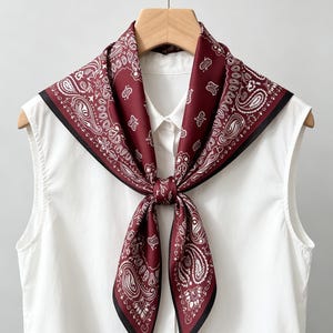 May include: A square scarf featuring a deep burgundy background with a white paisley pattern and a thin black border. The scarf is tied in a knot at the center and draped over a white sleeveless collared shirt hanging on a wooden hanger.
