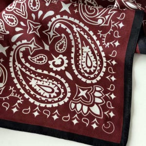 May include: A square fabric scarf featuring a deep burgundy background with a white paisley pattern. The design includes intricate teardrop shapes, star motifs, and floral elements. The scarf is finished with a solid black border along the edge.