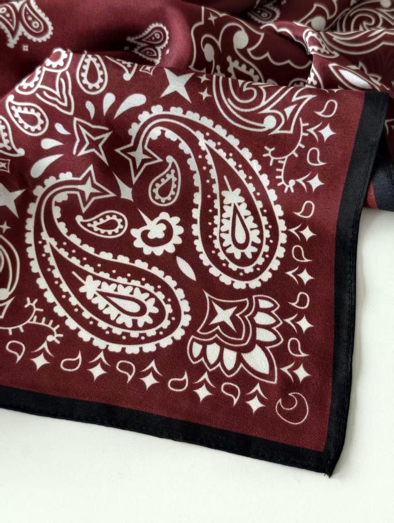 May include: A square fabric scarf featuring a deep burgundy background with a white paisley pattern. The design includes intricate teardrop shapes, star motifs, and floral elements. The scarf is finished with a solid black border along the edge.