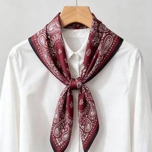 May include: A square scarf featuring a deep burgundy background with a white paisley pattern and a thin black border. The scarf is tied in a simple knot at the center and is draped over a crisp white collared button-down shirt on a wooden hanger.