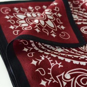 May include: A square bandana featuring a deep burgundy red base color with a contrasting white paisley pattern. The fabric has a smooth, slightly reflective finish and is bordered by a solid black hem that frames the intricate floral motifs.