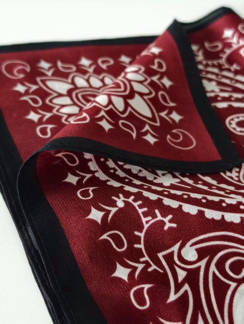 May include: A square bandana featuring a deep burgundy red base color with a contrasting white paisley pattern. The fabric has a smooth, slightly reflective finish and is bordered by a solid black hem that frames the intricate floral motifs.