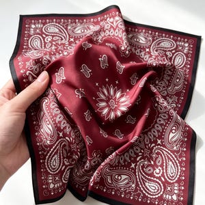 May include: A square scarf in deep burgundy with a white paisley pattern and a central floral motif. The fabric has a smooth, satin-like finish and is framed by a solid black border along all four edges, creating a distinct contrast in color.