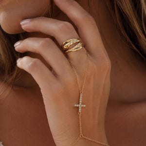 Op de afbeelding: Een goudkleurige handketting met een ring met een glad, golvend oppervlak, verbonden door een fijne ketting met een centrale kruishanger bezet met kleine, heldere, fonkelende steentjes op de rug van de hand.