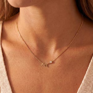 Puede incluir: Un delicado collar de oro con las iniciales "MH" y una pequeña gema brillante. El collar se muestra en el cuello de una persona, destacando su fina cadena y diseño minimalista. Un accesorio sutil pero elegante.