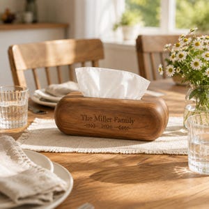 Puede incluir: Una caja de madera rectangular para pañuelos con bordes redondeados, grabada con el texto "The Miller Family" sobre el año "2026" y detalles de hojas. Un pañuelo blanco sobresale por la abertura superior del soporte de madera marrón.
