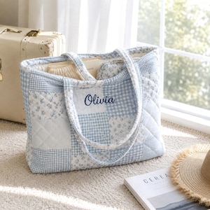 May include: A quilted tote bag featuring a patchwork design of light blue and white gingham checks and floral patterns. The bag has two shoulder straps and the name Olivia embroidered in dark blue script on the front center panel.
