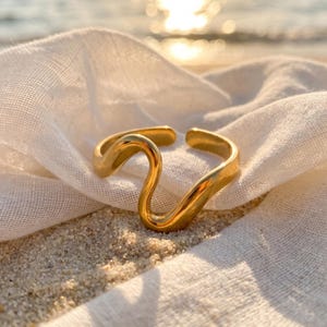 Può includere: Un anello in metallo dorato caratterizzato da un design aperto, ondulato e fluido. La fascia presenta una superficie liscia e lucida che si curva in una distinta forma a S, creando un'apertura regolabile sulla parte superiore. Il metallo brilla.
