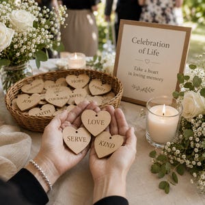 Könnte beinhalten: Drei kleine herzförmige Holzanhänger mit den eingravierten Wörtern LOVE, SERVE und KIND. Die Anhänger werden vor einem geflochtenen Korb mit weiteren Holzherzen, einem Schild mit der Aufschrift Celebration of Life und einer Kerze gehalten.