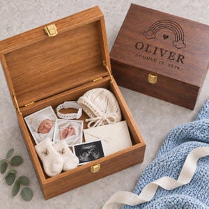 May include: Two wooden keepsake boxes with gold latches. One box is open, containing a white knit baby hat, a white hospital bracelet, two baby photos, a pair of cream knit booties with bear faces, and a white envelope. The closed box features a rainbow engraving with the name OLIVER and the date AUGUST 12, 2026.