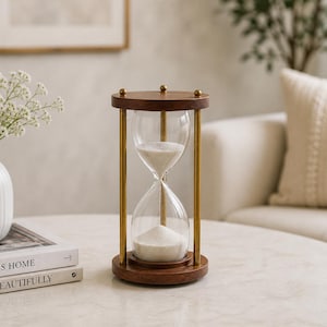 May include: An hourglass with a wooden top and base, brass-colored supports, and clear glass containing white sand. The hourglass is on a marble-topped table, with a vase of white flowers and books nearby. The decor has a neutral color palette.