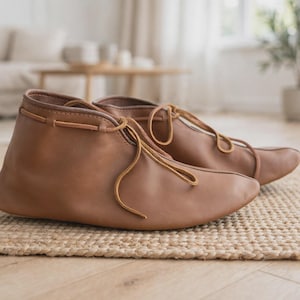 Op de afbeelding: Een paar enkelhoge laarzen gemaakt van glad, lichtbruin leer. Deze laarzen hebben een afgeronde neus en worden vastgemaakt met dunne, bijpassende bruine leren veters die door de bovenste rand lopen en aan de voorzijde in een strik zitten.