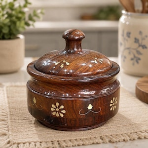 Puede incluir: Un recipiente redondo de madera de color marrón oscuro con una tapa a juego y un pomo central. El exterior del recipiente y la tapa están decorados con pequeños motivos florales de latón incrustados y delicadas líneas curvas en secuencia.