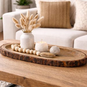 Puede incluir: Una mesa de centro de madera con una bandeja decorativa que contiene un jarrón blanco con flores secas, una vela, cuentas de madera y una esfera de piedra. La bandeja tiene un borde de corteza natural. La mesa está frente a un sofá blanco.