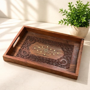 May include: A rectangular wooden serving tray featuring intricate floral carvings along the border and a central inlay of small brass leaf-shaped accents. The tray has two integrated side handles and a dark brown finish with visible wood grain.