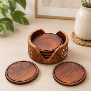 May include: A set of circular wooden coasters with a dark brown grain and etched floral patterns, housed in a matching cylindrical wooden holder featuring white inlaid leaf designs on the exterior, with two coasters placed in front.