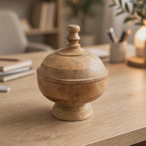 Puede incluir: Un cuenco de madera de color marrón claro con tapa, cuerpo redondeado y base de pedestal. La tapa presenta un remate central con un diseño torneado de varios niveles. La veta natural de la madera es visible en toda la superficie.