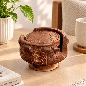 May include: A set of round wooden coasters stacked inside a matching carved wooden holder. The coasters and the holder feature a dark brown finish with intricate, hand-carved floral or sunburst patterns on the top surfaces and sides.