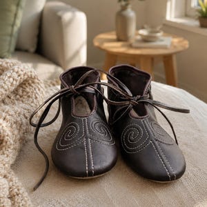 May include: A pair of dark brown leather shoes featuring a rounded toe, a high-cut ankle, and thin leather laces tied at the front. The exterior surface of each shoe is decorated with white stitched spiral patterns and vertical lines.