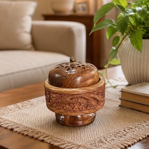 Peut inclure: Un récipient rond en bois avec un motif floral sculpté sur le côté et une finition polie lisse. Il comporte un couvercle bombé amovible avec un petit bouton en laiton sur le dessus et des trous de ventilation en forme d'étoile découpés.