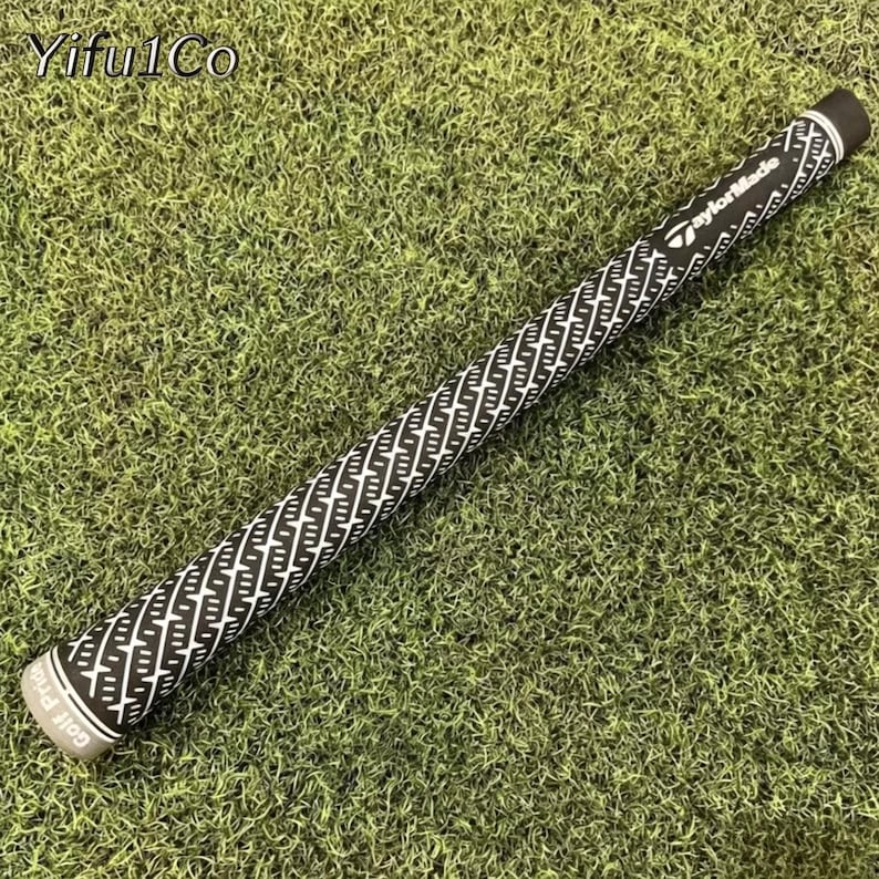 Puede incluir: Un grip de palo de golf en blanco y negro con un patr&oacute;n geom&eacute;trico repetitivo. El grip tiene el texto "TaylorMade" y "Golf Pride" impreso. El grip est&aacute; sobre un lecho de hierba verde.