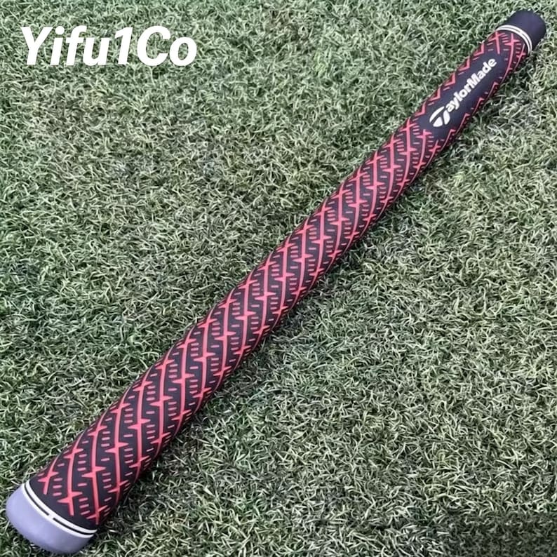 Puede incluir: Un grip de palo de golf con un patr&oacute;n geom&eacute;trico negro y rojo. El grip tiene una tapa gris con rayas blancas y el logotipo de TaylorMade. El grip est&aacute; sobre una superficie verde.
