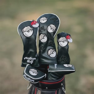 Könnte beinhalten: Ein Satz schwarzer Golfschlägerhauben mit weißer Umrandung und einem Golfball-Design mit Hut. Die Hauben sind in einer Golftasche angeordnet, mit dem Logo "Malbon Golf" auf einigen.