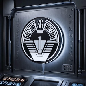 May include: A circular black and white emblem with the letters "SG" and the number "1" on a metallic panel. The emblem is surrounded by a white light, and the panel has additional symbols and the letters "SGC".